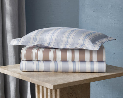 Striped Elio sheets in tan and blue, stacked on a stone side table.