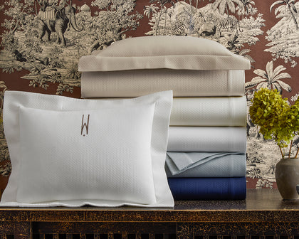 White Elliot textured pique bedding stacked in ivory, gray, and navy, beside a monogrammed pillow.