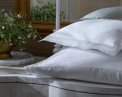 White Grace pillows with lace trimmed edges on a bed near a mirror and flowers.
