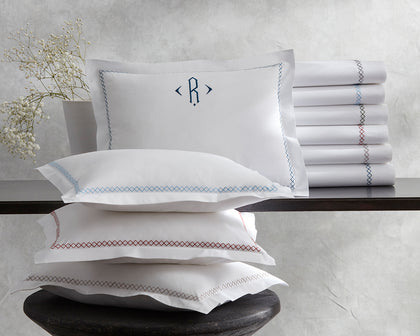 White Hatch pillows and rolled sheets with cross-stitch trim in assorted colors, beside a monogrammed sham and baby's breath.