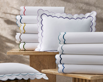 White India scalloped sheets and shams stacked on stone tables in a rainbow of trim colors.