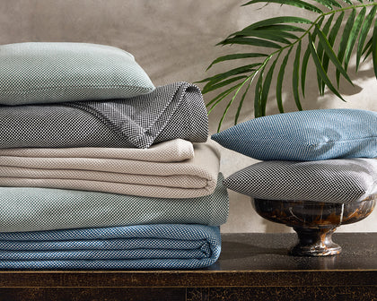 Stacked Jackson houndstooth-textured sheets and pillows in sage, black, tan, and blue, beside a palm frond.