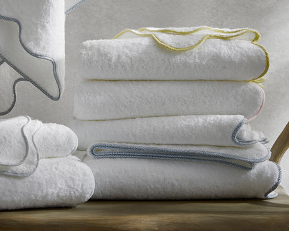 White Jasper towels with braided trim in yellow, gray, and pink, stacked on a wood bench.