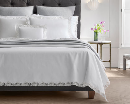 White Lace Pique bedding with floral lace trim, styled on a bed with a gray tufted headboard.