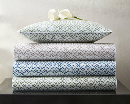 Laksha medallion print sheets stacked in green, tan, blue, and olive, topped with a calla lily.
