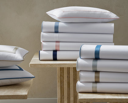 White Louise sheets and pillows stacked with satin bordered trim in assorted colors.