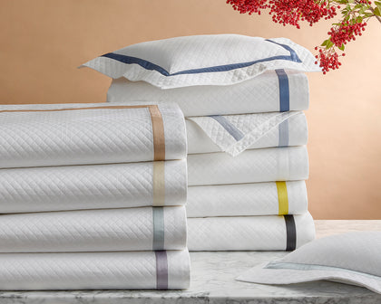 White Lowell Matelasse quilted shams stacked with ribbon trim in assorted colors, beside red berries.
