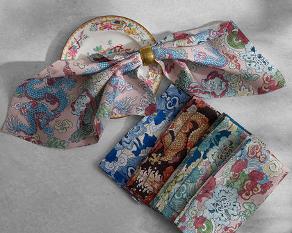 Magic Mountain dragon print napkins in pink, blue, and gold, folded with a napkin ring beside a floral plate.