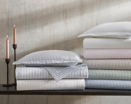 Blue-striped Matteo bedding folded on a dark shelf beside lit taper candles, with striped bedding in assorted colors stacked alongside.