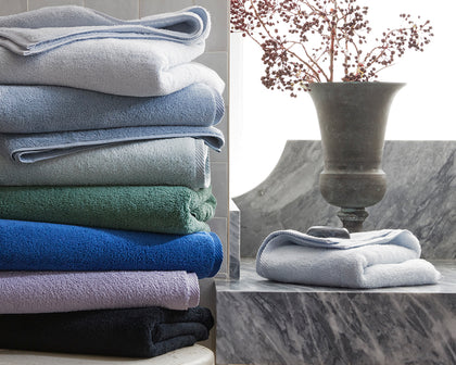 Milagro bath towels stacked in assorted colors beside a folded towel on marble with a dried berry arrangement.