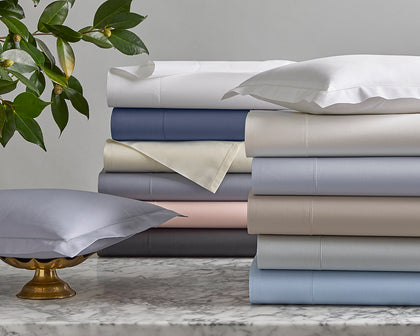 Milano Hemstitch sheets stacked in assorted colors on marble beside a lavender pillow in a brass stand.