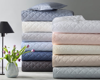 Milano Quilt bedding stacked in assorted colors beside a marble vase of tulips and a black lamp.