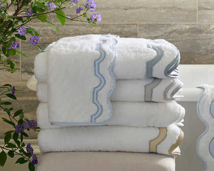 White Mirasol towels with scalloped blue, gray, and gold trim stacked beside purple flowers.