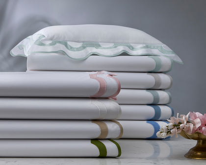 White Mirasol bedding with scalloped multicolor trim stacked on marble beside pink flowers.
