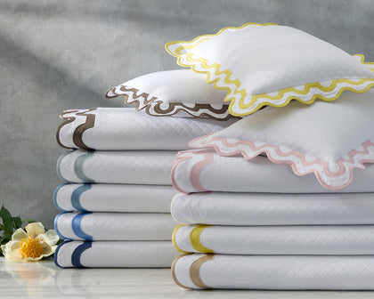 White Mirasol Matelasse coverlets with scalloped multicolor trim stacked with shams beside a white flower.