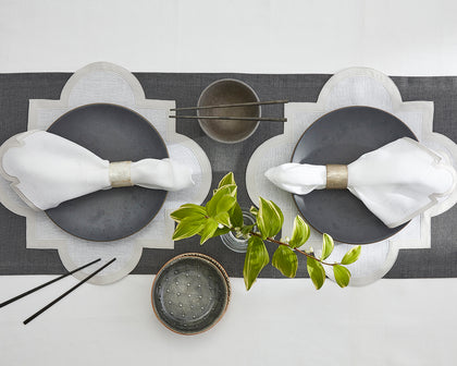 Gray scalloped-edge placemats set on a table runner with dark plates and greenery.