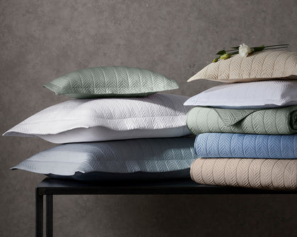 Netto herringbone-quilted shams and bedding stacked in assorted colors on a black console.