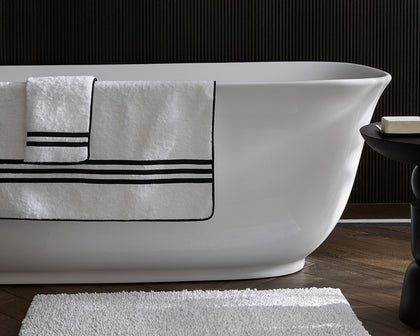 White Newport towel with black striped border draped on a freestanding tub, bath mat below.