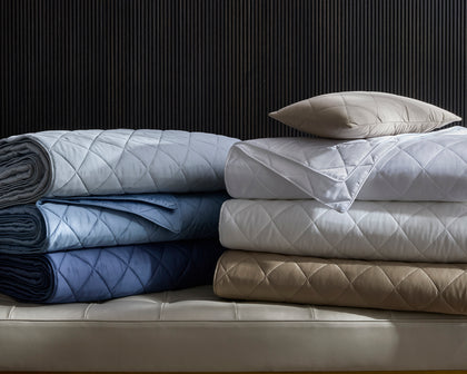Diamond-quilted Nocturne Quilt bedding stacked in assorted colors on a quilted bench.