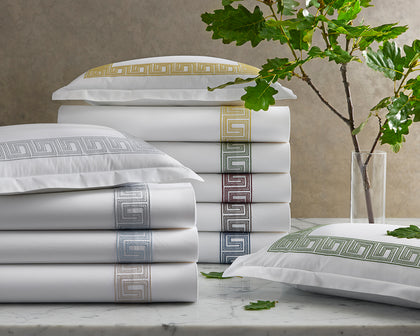 White Ophelia sheets with Greek key embroidered trim in assorted colors stacked on marble with oak leaves.