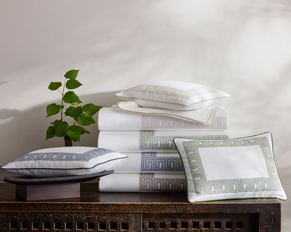 White Ophelia Pique bedding with Greek key trim in assorted colors stacked on a carved wood console.