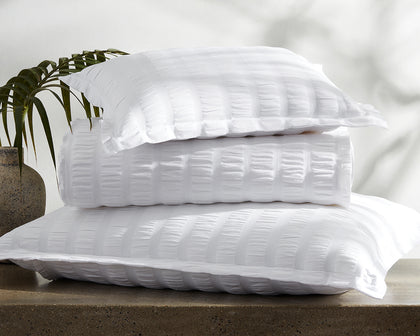 White seersucker-textured Panama bedding stacked on a stone ledge beside a palm frond.