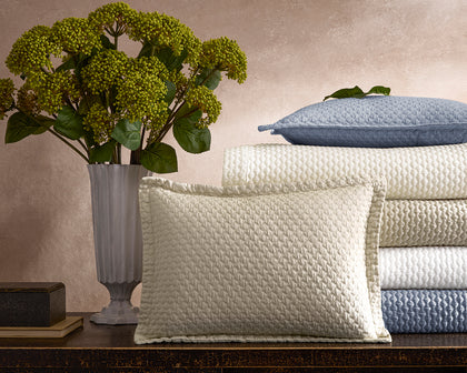 Textured Perla quilted bedding stacked in cream and blue on a console beside hydrangeas.