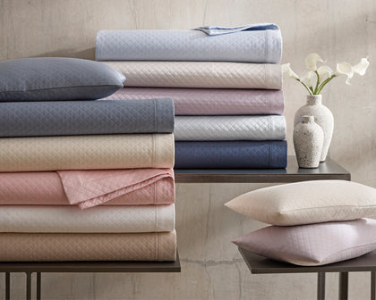 Diamond-quilted Petra bedding stacked in assorted colors on a table beside white ceramic vases.