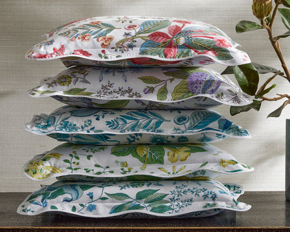 White Pomegranate floral-print pillows in assorted colorways stacked beside a leafy plant.