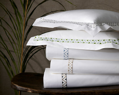 White Prado sheets with zigzag embroidered trim in assorted colors stacked on a wood table.
