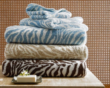 Zebra-print Regine towels in blue, taupe, and brown stacked on a stone ledge with a soap dish.