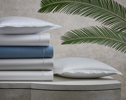 Talita Satin Stitch sheets stacked in white, blue, and silver on a stone bench beside palm fronds.