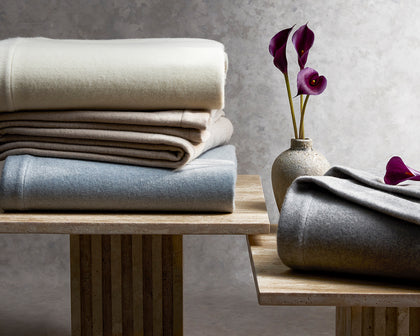 Venus wool blankets folded in assorted colors on wood stands beside purple calla lilies.
