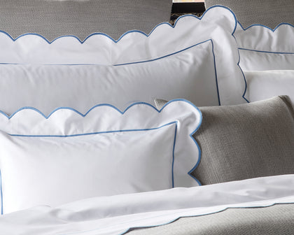 White Butterfield scalloped shams with blue trim, styled on a bed with a gray textured pillow.