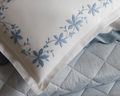 Blue Callista floral vine embroidered pillow, shown in detail on a speckled quilted bed.