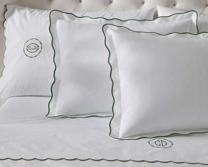 White Camilla Pique scalloped pillows with green trim and monograms, styled on a tufted bed.