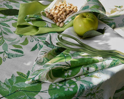 Green Citrus Garden leaf-print tablecloth and napkins set with a green apple and pistachios.