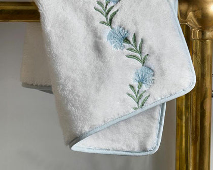 White Daphne towel with blue floral vine embroidery, hanging on a brass rack.