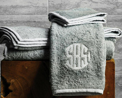 Gray Enzo towels with white trim and a monogram folded on a wooden stool.