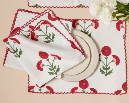 White Gisele placemats and napkins with embroidered red poppies, stacked with a gold rimmed plate.