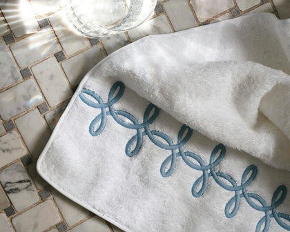 White Gordian Knot towel with blue embroidered knot trim on a marble tile floor.
