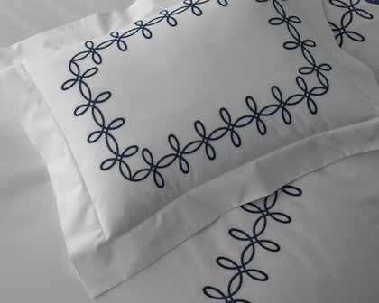White Gordian Knot pillow with a navy embroidered border on a bed.