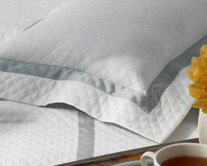 White Lowell Matelasse quilted pillow with gray ribbon trim, styled on a bed with a tray and yellow flower.