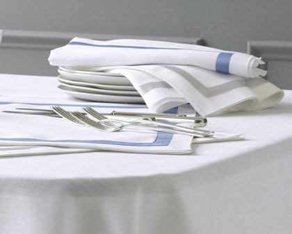 White Lowell napkins with blue and gray bordered trim, stacked with white plates and silver flatware.