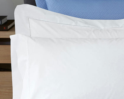 White Luca Hemstitch pillows with a hemstitch trim, styled on a bed with a blue quilted headboard.