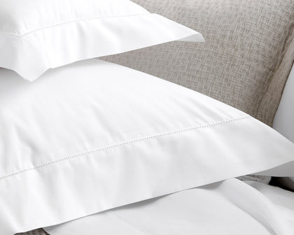 White Milano Hemstitch pillowcases and sheets styled on a bed with a woven headboard.