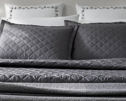 Charcoal diamond-quilted Milano Quilt shams and coverlet styled on a bed with vine-embroidered white pillowcases.