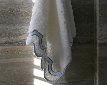 White Mirasol towel with scalloped blue trim hanging against a marble wall.