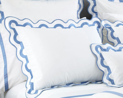 White Mirasol pillow shams with scalloped blue trim stacked on a bed.