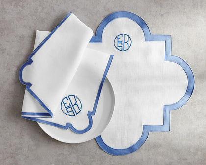 White scalloped Mirasol placemat with blue trim and monogrammed napkins on a plate.